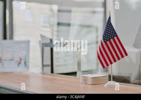 Close-up of American flag standing on office desk with sunlight filtering through blinds. Background including few office supplies and decor items cre Stock Photo