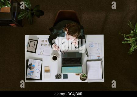 Man analyzing charts and graphs at desk with laptop, planning business strategy. Surrounded by office supplies and looking at project details with foc Stock Photo