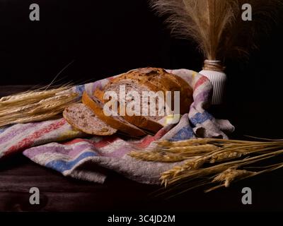 Sliced rustic bread on a colorful kitchen towel with wheat ears and a decorative vase on a dark wooden background Stock Photo