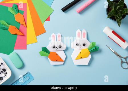 White handmade origami bunnies surrounded by art and hobby supplies: colored paper, glue, eraser, scissors. Top view Stock Photo
