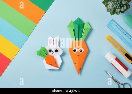 White origami rabbit and orange paper carrot on work table with tools for creativity and hobby: colored paper, glue, ruler, scissors. Top view Stock Photo