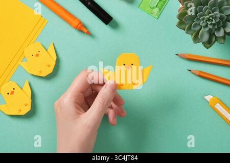 Making a simple Easter origami handicraft. Holds in his hand a ready-made paper craft of a chicken. Around the tools for crafts Stock Photo