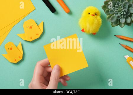 Making a simple Easter origami handicraft. The hand holds a square piece of yellow paper. Around the ready-made crafts chickens and tools for crafts Stock Photo