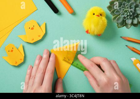 Making a simple Easter origami handicraft. Hands fold a square yellow sheet in half with a ruler. Around ready-made crafts chickens and tools for craf Stock Photo