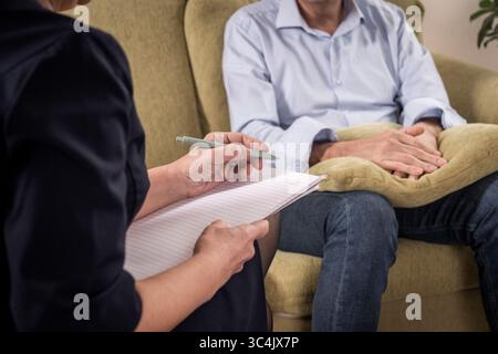 Close up on a therapist taking notes while her male patient holds a cushion in his lap for comfort and security during a difficult counseling session. Stock Photo