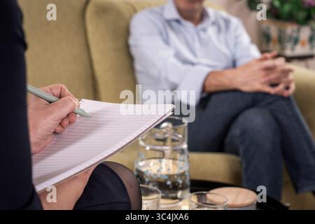 Over the shoulder view of a female psychologist or counselor writing in a notepad while listening to a mature male patient during a therapy session. Stock Photo