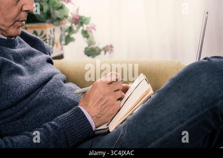 Anonymous close-up of a mature man's hands holding a pen and writing thoughts or taking notes in a diary during a remote work call or study session. Stock Photo