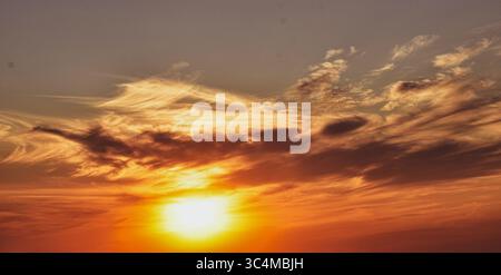 Fiery clouds float across the twilight sky as the golden sun sets gently behind the distant horizon. Stock Photo