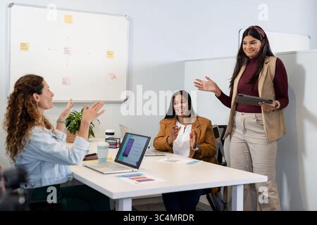 Diverse female coworkers collaborating at office table using laptops tablet charts coffee cup plant Stock Photo