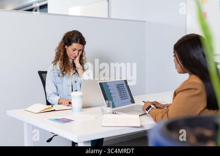 Diverse female coworkers wearing professional attire working on project at office desk with laptops Stock Photo