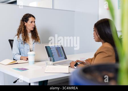 Diverse female colleagues discussing project details at office table using laptops and notebooks Stock Photo