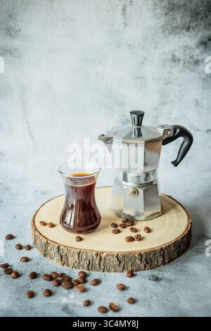 Curved glass of hot coffee on coaster with scattered beans, bowl of roasted beans and blurred moka pot on tree-slice board behind, spacious backdrop f Stock Photo