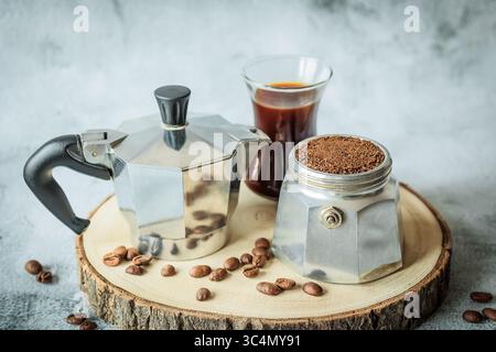 Stovetop moka pot, basket packed with ground coffee and freshly brewed cup arranged on a rustic wood slice, scattered beans over light concrete, cozy Stock Photo