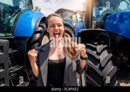 Successful businesswoman celebrating with clenched fists near tractors at agricultural equipment dealership Stock Photo