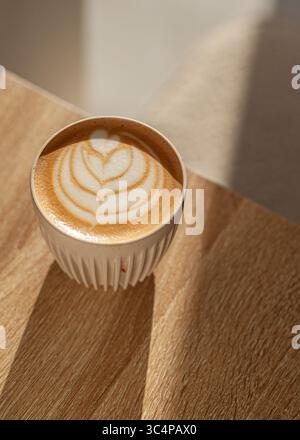 A ceramic cup of cappuccino with elegant cream latte art sits on a light wooden table in a minimalist coffee shop setting. Stock Photo