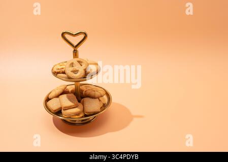 A golden two-tier cookie stand with Danish butter cookies captured from a slight angle. The heart-shaped handle and peach-toned background give a cozy Stock Photo