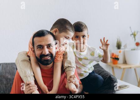 Happy middle eastern father having fun with his three sons at home. Dad cuddling toddler while other kids play and smile. Loving family moment on cozy Stock Photo