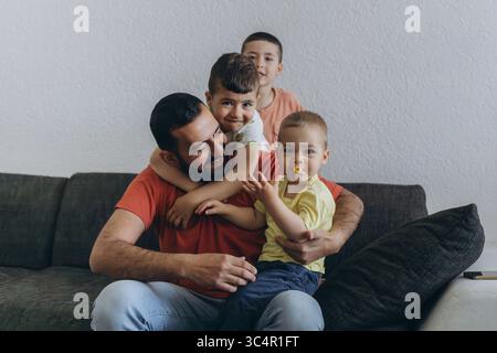 Happy middle eastern father having fun with his three sons at home. Dad cuddling toddler while other kids play and smile. Loving family moment on cozy Stock Photo