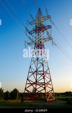 A tall red and white lattice transmission tower carries high voltage power lines across a rural landscape at sunset. Stock Photo