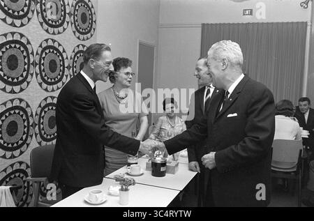 Congratulations to the anniversary Whizgle News, Dutch Desk, The Netherlands, 1950-2000 A group of people are gathered in a well-decorated room, engaged in conversation and exchanging greetings. Two men are shaking hands, while a woman stands nearby, smiling. In the background, additional attendees participate in the lively atmosphere, with coffee cups placed on tables. The setting suggests a formal or celebratory event. Stock Photo