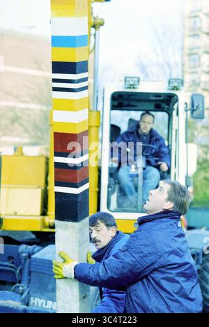 Installing artwork, Schalkwijk, Haarlem, The Netherlands, 11-12-1995 Whizgle News, Dutch Desk, The Netherlands, 1950-2000 Two workers in blue jackets are positioned near a colorful striped pole, focusing intently on their tasks. Behind them, a vehicle operates, with an operator visible in the cab. The scene conveys a sense of teamwork and the integration of construction-related activities. Stock Photo