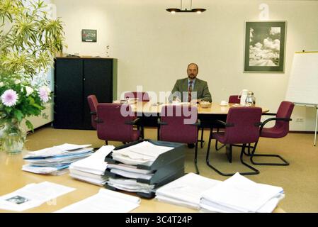 Mr. Bongers, Nova College, 11-04-1999 Whizgle News, Dutch Desk, The Netherlands, 1950-2000 A man in formal attire sits alone at a round conference table surrounded by chairs in a minimalistic office space. Papers and documents are stacked on a desk nearby, and a flowering plant adds a touch of nature to the room. A whiteboard and framed photographs are visible on the walls. Stock Photo