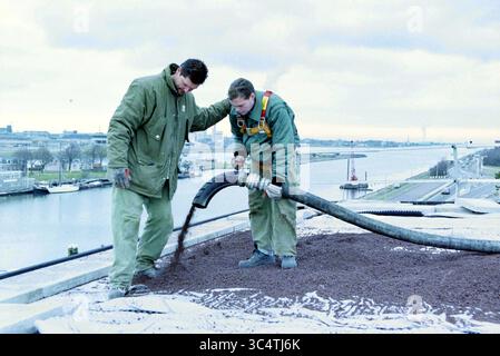 Spraying lava rock on the roof of Rijkswaterstaat, IJmuiden, IJmuiden, The Netherlands, 17-02-1999 Whizgle News, Dutch Desk, The Netherlands, 1950-2000 Two workers are engaged in a construction task on a rooftop, carefully operating a hose that dispenses material onto the surface. The backdrop features a river and distant buildings, hinting at an urban setting. Both individuals are dressed in protective gear, showcasing their focus and teamwork in this industrial environment. Stock Photo