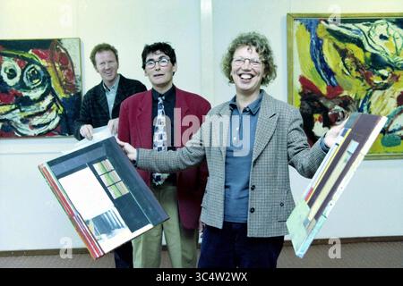 Presentation of the public award from the municipality of Haarlem for the works of Helmuth van Galen (with blue checked jacket)., Haarlem, The Netherlands, 11-04-1999 Whizgle News, Dutch Desk, The Netherlands, 1950-2000 Three individuals stand together in an art gallery, showcasing their enthusiasm for creative expression. Two men wear distinctively patterned jackets, while a woman proudly holds up vibrant paintings, smiling broadly. Behind them, colorful abstract artworks adorn the walls, enhancing the lively atmosphere of the space. Stock Photo