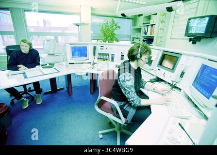 Haarlem ambulance service control room, Haarlem, The Netherlands, 22-02-2000 Whizgle News, Dutch Desk, The Netherlands, 1950-2000 A workspace featuring two individuals focused on their tasks. One person is seated at a computer, wearing a headset and typing, while the other is sitting nearby, looking at a screen. The area is equipped with multiple monitors and electronics, creating a dynamic office environment. A plant adds a touch of greenery to the setting. Stock Photo