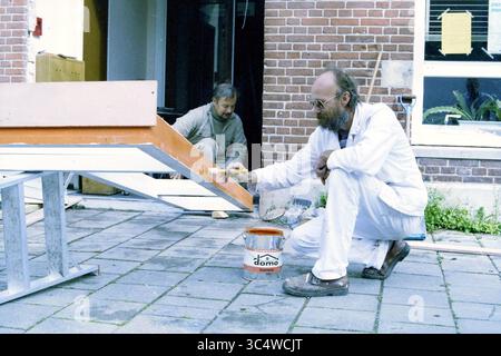 Construction worker and painters, Employment Board Kennemerland, Activities, 16-09-1988 Whizgle News, Dutch Desk, The Netherlands, 1950-2000 Two men are engaged in a painting project outdoors. One man is kneeling and applying orange paint to a large wooden structure, while the other man is working in the background, focused on a different task. The setting features a brick wall and a work area filled with tools and materials. Stock Photo