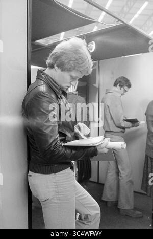 Job market opening of Minister Albeda and stands Kennemersporthal, Market, Politics, people politics, Haarlem, The Netherlands, 23-08-1979 Whizgle News, Dutch Desk, The Netherlands, 1950-2000 A young man in a leather jacket leans against a wall, intently reading a document while holding a rolled-up paper. In the background, another individual appears to be reviewing something in a more relaxed manner, creating a sense of a busy, focused environment. Stock Photo