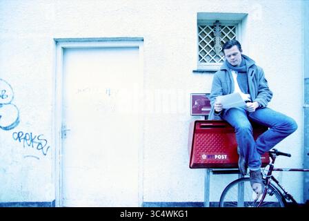 Pim Bijtel, Haarlem, The Netherlands, 06-03-2001 Whizgle News, Dutch Desk, The Netherlands, 1950-2000 A person sits casually on a bicycle, leaning against a red mailbox while reviewing some papers. The backdrop features a plain wall and a closed door, with subtle graffiti adding a touch of urban character to the scene. Stock Photo