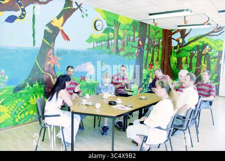Mural TPG, Haarlem, The Netherlands, 12-06-2003 Whizgle News, Dutch Desk, The Netherlands, 1950-2000 A diverse group of individuals gathers around a table, sharing a meal and conversation in a vibrant, nature-themed room adorned with colorful murals of trees and wildlife. The atmosphere is relaxed and communal, with each person engaged in their surroundings. Stock Photo