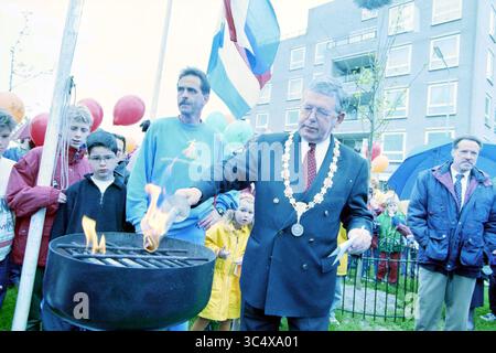 Mayor of Houwlingen lights BBQ at a party, Haarlemmermeer, 04-05-1998 Whizgle News, Dutch Desk, The Netherlands, 1950-2000 A group of people gathers at an outdoor event, with balloons in the background. A man in formal attire, wearing a ceremonial necklace, lights a fire in a grill while onlookers, including children, watch with interest. The atmosphere appears festive and communal, suggesting a celebration or special occasion. Stock Photo