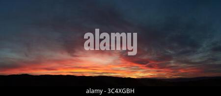 Stunning sunset paints sky with vibrant hues of red, orange, and pink, blending into deep blues and purples. Silhouetted hills create contrast against fiery horizon, adding drama to serene landscape. Stock Photo