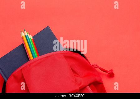 A red backpack with a notebook and colorful pencils on red background. Back to school concept with copy space. Stock Photo