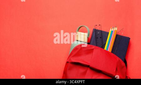 A red backpack with a notebook, a ruler, a square, colorful pencils and a bottle on red background. Back to school concept with copy space. Stock Photo