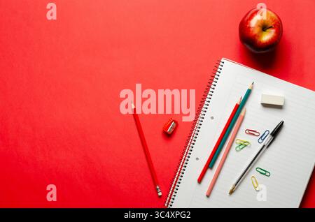 An apple, a notebook, some colorful pencils, a pen, an eraser, a sharpener, and paperclips on red background and copy space. Back to school concept. Stock Photo