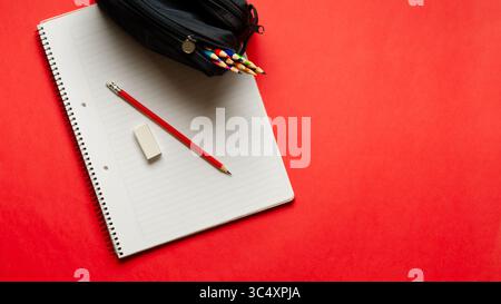 A notebook with a pencil and an eraser, and a pencil case full of colorful pencils on red background and copy space. Back to school concept. Stock Photo