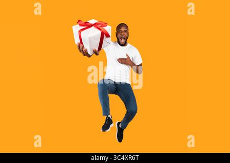 Funny Black Guy Jumping Holding Gift Box Over Yellow Background Stock Photo