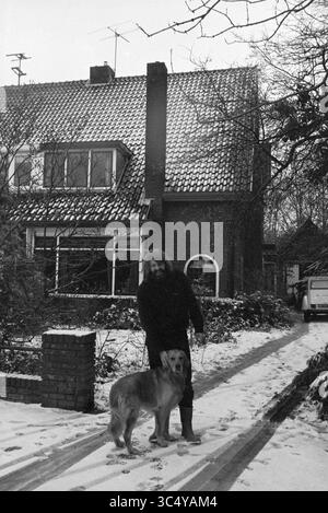 Man with dog in front of large corner house, 15-03-1979 Whizgle News, Dutch Desk, The Netherlands, 1950-2000 A person stands beside a golden retriever on a snow-covered path, with a cozy house featuring a tiled roof in the background. The scene captures a peaceful winter moment, surrounded by trees and a quiet neighborhood atmosphere. Stock Photo