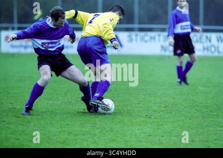 Football: @@ - VZW, 16-03-2001 Whizgle News, Dutch Desk, The Netherlands, 1950-2000 Two soccer players compete for the ball on a grassy field. One player, wearing a purple jersey, attempts to gain control as the other, dressed in yellow and blue, skillfully maneuvers to evade the challenge. In the background, additional players are positioned, ready to engage in the match. Stock Photo