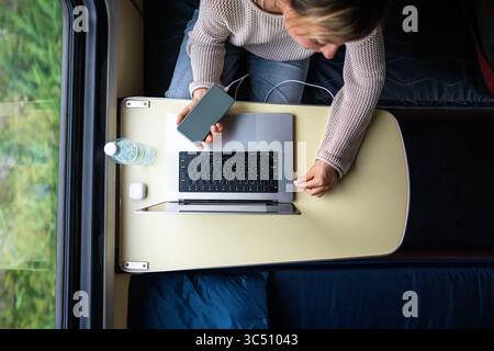 Woman plugging laptop into power bank, charging batteries, sitting in train compartment. Stock Photo