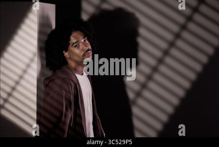 Fashionable young african-american man with curly hair posing in studio near wall with striped shadows Stock Photo