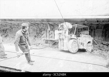 Unloading 'Bretagne', Wijsmuller, 15-04-1966 Whizgle News, Dutch Desk, The Netherlands, 1950-2000 A worker operates a large industrial vehicle, maneuvering it carefully within a confined space, while another individual stands nearby, holding a chain attached to the machinery. The environment appears rugged and gritty, typical of a construction or excavation site, with debris scattered around. Stock Photo