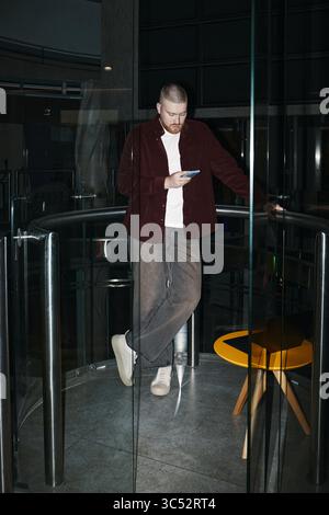Caucasian young man standing indoors, leaning against glass railing, using smartphone with focused expression, modern interior visible, in coworking office Stock Photo