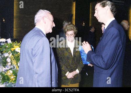 Group and reception Ven, 06-11-1990 Whizgle News, Dutch Desk, The Netherlands, 1950-2000 A group of three individuals engages in a conversation at a formal event, with two men and a woman present. The man on the left smiles as he interacts with the woman in a patterned outfit, who appears contemplative. The other man, dressed in a suit, holds a small blue gift box while listening attentively. Floral arrangements decorate the setting, adding a touch of elegance to the scene. Stock Photo