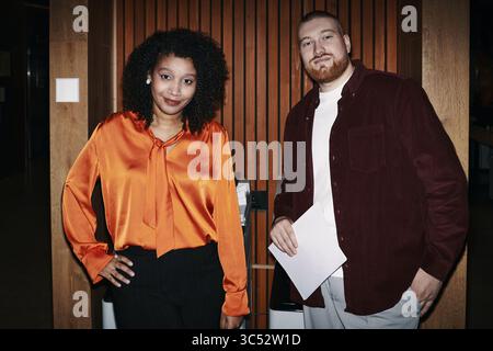 Young Black woman and Caucasian man standing together in office, woman resting hand on hip while man holding document, both colleagues looking confidently at camera Stock Photo
