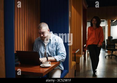 Caucasian young male entrepreneur working on laptop at table in modern workspace, focusing on screen while Black young adult woman walking in background, both engaged in professional activities Stock Photo