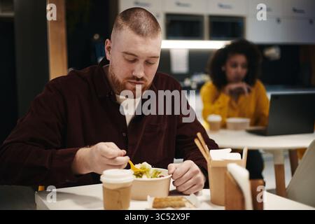 Caucasian young man eating salad at table in modern cafe, holding fork and looking down at food, takeaway coffee cup and disposable container in front, Black woman working on laptop in background Stock Photo